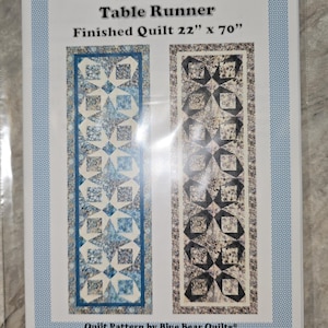 Puede incluir: Un patrón de acolchado para un camino de mesa llamado "Periwinkle Prisms". El patrón es para una colcha terminada que mide 56 cm por 178 cm. El patrón incluye dos diseños diferentes, uno en azul y blanco y otro en negro y blanco. El patrón es de Blue Bear Quilts y tiene derechos de autor de 2021.