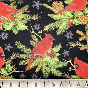 May include: A black fabric featuring a repeating pattern of bright red cardinal birds perched on green pine branches with red berries, pinecones, and grey snowflakes. A white measuring tape is visible at the bottom, showing 15 inches in length.