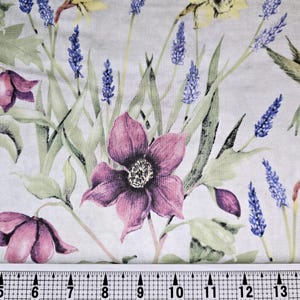 May include: A white fabric with a floral print featuring purple flowers, green leaves, and a hummingbird. The fabric has a ruler with inches marked on it.