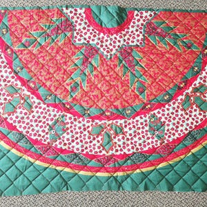 May include: A quilted Christmas tree skirt with a festive design. The skirt features a red and white patterned center with green and red accents. Two square starburst designs are on the corners. The skirt is approximately 60 inches wide.