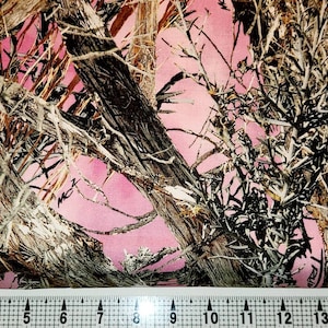 May include: Pink camouflage fabric with a pattern of brown and gray tree branches. A ruler is at the bottom of the image, showing measurements in inches. The fabric has a pink background.