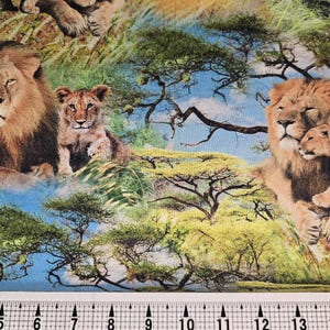 Könnte beinhalten: Stoff mit einem lebendigen Druck mit Löwen und Jungtieren in einer Savannenlandschaft. Das Design umfasst üppiges grünes Gras, Bäume und einen blauen Himmel und schafft eine realistische Wildtierszene. Das Lineal unten misst bis zu 40 cm.