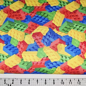 May include: Fabric featuring a repeating pattern of toy building blocks in red, yellow, green, and blue. The blocks are scattered across the fabric, creating a playful design. A ruler is at the bottom, showing measurements.