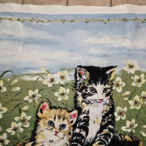 May include: A fabric panel featuring two kittens in a field of white flowers. One kitten is black and white, the other is tan and brown. The background shows a blue sky with clouds and a decorative border.