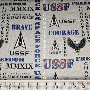 May include: Gray fabric with a repeating pattern featuring the USSF logo, eagles, and the words "Freedom," "Courage," and "Brave." The text "United States Space Force" is also visible. The fabric has a ruler at the bottom.