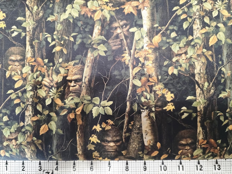May include: A fabric design depicting a forest scene with multiple Bigfoot figures concealed within the trees and leaves. The colour scheme is dominated by greens, browns, and yellows, creating a camouflage effect. The Bigfoot figures have detailed facial features and fur.