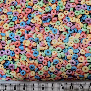 Puede incluir: Una imagen de primer plano de una tela con un patrón de anillos de cereal coloridos. La tela está impresa con una variedad de colores, incluyendo rojo, naranja, amarillo, verde, azul y morado. Los anillos de cereal están dispuestos en un patrón aleatorio, creando un diseño divertido y juguetón.