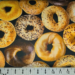 May include: A close-up of a fabric with a repeating pattern of various types of bagels. The bagels are a mix of light and dark brown with different toppings, including sesame seeds, poppy seeds, and everything seasoning.