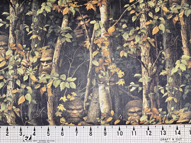 May include: A forest-themed fabric featuring multiple Bigfoot figures hidden amongst trees and leaves. The fabric's background is dark, with green, brown, and yellow hues. A ruler at the bottom displays measurements in inches.