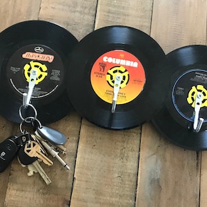 May include: Three black vinyl records with yellow and white labels are mounted on a wall with hooks. Each record has a silver hook attached to the center label. A set of keys is hanging from one of the hooks.
