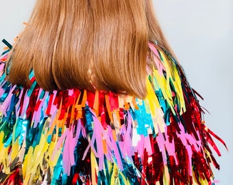 May include: A colorful, fringed garment with a variety of bright colors, including red, yellow, pink, and blue. The fringe is made of shiny, metallic material. The person's hair is long and light brown.