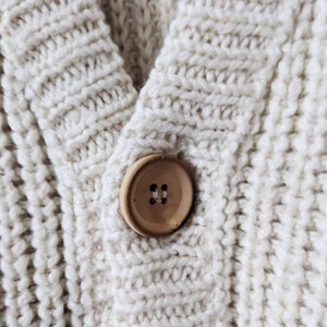 May include: A close-up of a cream-colored knitted cardigan with a wooden button. The button has two holes.