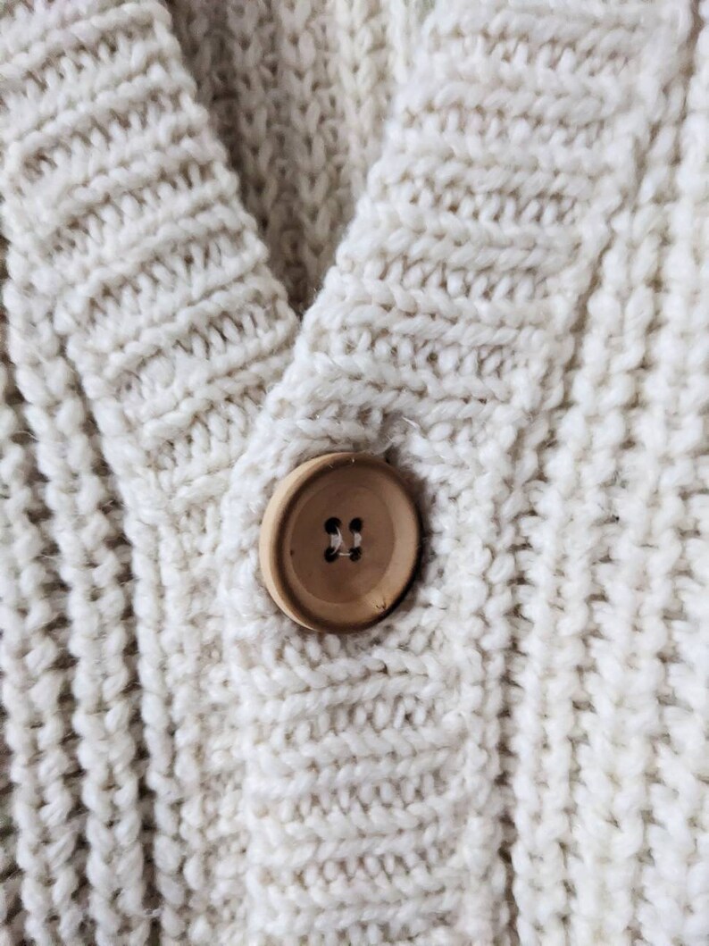May include: A close-up of a cream-colored knitted cardigan with a wooden button. The button has two holes.