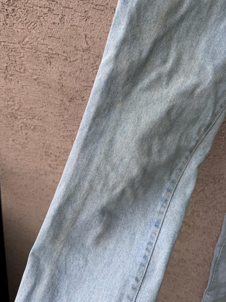 K&ouml;nnte beinhalten: Nahaufnahme von hellblauem Denim-Stoff. Der Denim hat ein strukturiertes Aussehen mit subtilen Farbvariationen und sichtbaren N&auml;hten entlang der N&auml;hte. Der Stoff ist leicht zerknittert, was darauf hindeutet, dass er getragen oder gewaschen wurde. Der Gesamteindruck ist l&auml;ssig und klassisch.
