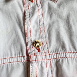 May include: A white button-down shirt featuring prominent orange contrast stitching along the placket and horizontal seams. The garment includes two visible gold-toned metallic buttons with embossed detailing, each secured with orange thread.