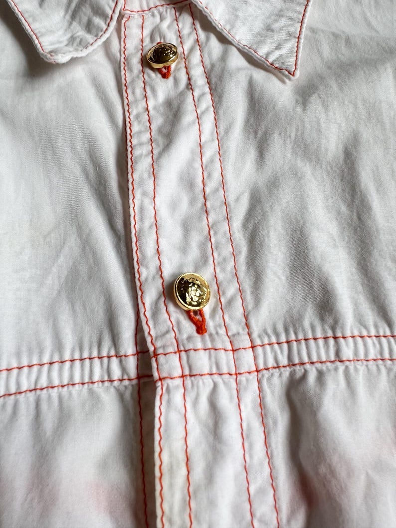May include: A white button-down shirt featuring prominent orange contrast stitching along the placket and horizontal seams. The garment includes two visible gold-toned metallic buttons with embossed detailing, each secured with orange thread.