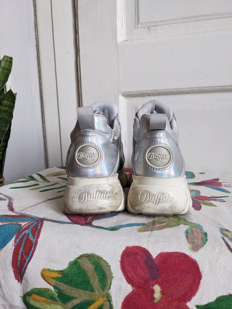 Op de afbeelding: Een paar zilveren platform sneakers met het woord "Buffalo" gedrukt op de zijkant en achterkant van elke schoen.