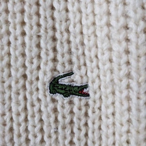 May include: White knitted sweater with a green crocodile logo embroidered on the front.