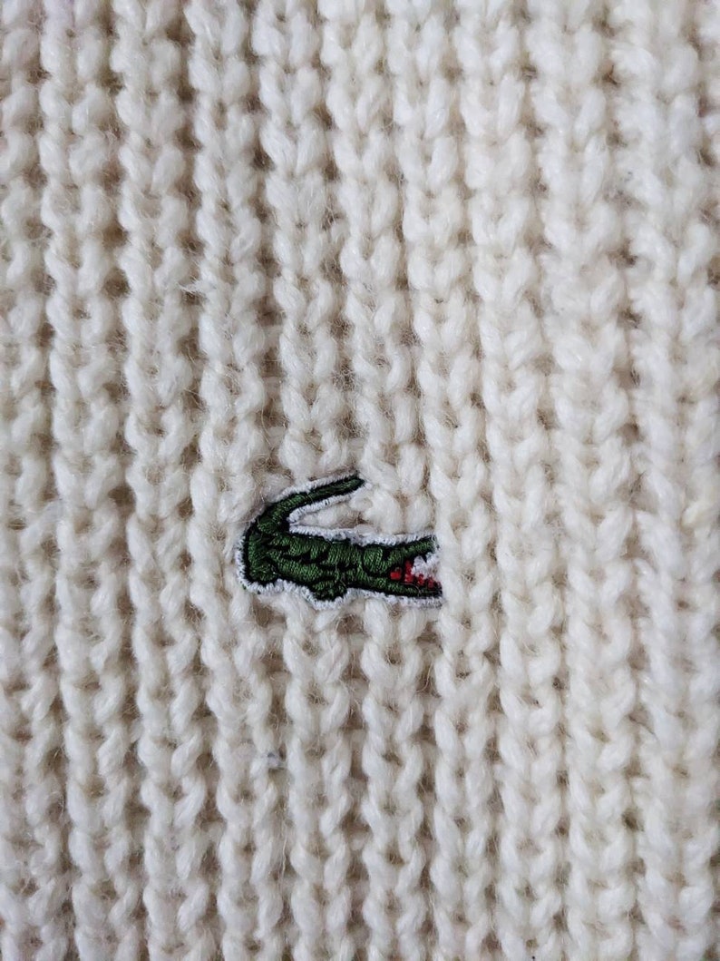 May include: White knitted sweater with a green crocodile logo embroidered on the front.