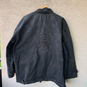Puede incluir: Chaqueta vaquera negra con el logotipo de Versace Jeans Couture bordado en la espalda. La chaqueta tiene un cuello cl&aacute;sico, cierre de botones y dos bolsillos en el pecho. La chaqueta est&aacute; colgada en una percha de madera.
