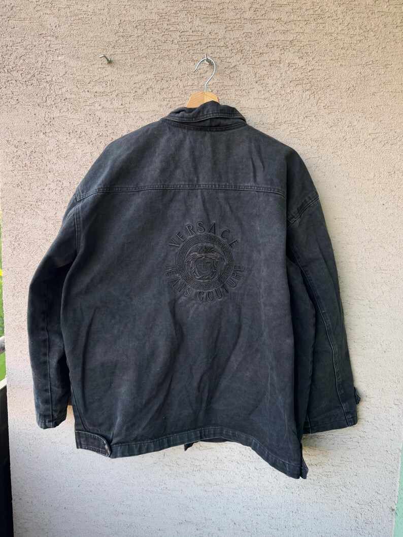 Puede incluir: Chaqueta vaquera negra con el logotipo de Versace Jeans Couture bordado en la espalda. La chaqueta tiene un cuello cl&aacute;sico, cierre de botones y dos bolsillos en el pecho. La chaqueta est&aacute; colgada en una percha de madera.