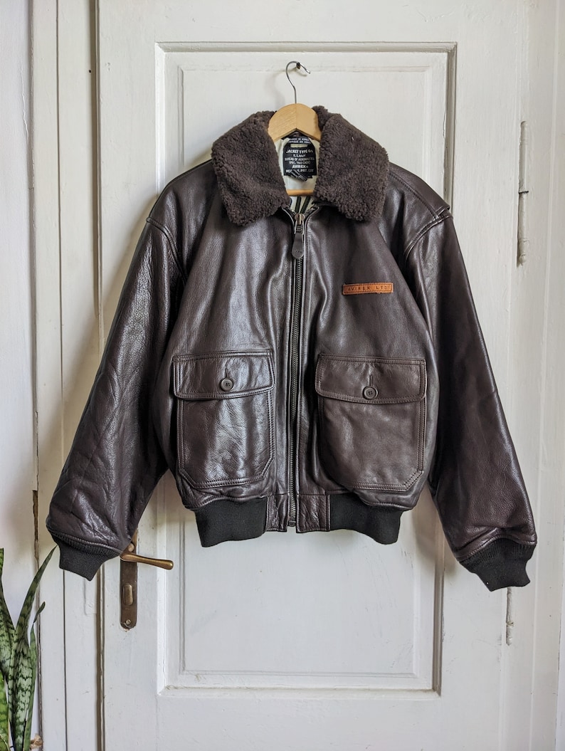 May include: A brown leather bomber jacket with a shearling collar, two front pockets, and a black ribbed hem and cuffs. The jacket has a zipper closure and a small patch that reads "AVIREX NYC". The jacket is hanging on a wooden hanger.
