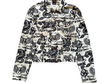 Könnte beinhalten: Eine cremefarbene Jeansjacke mit einem durchgehenden schwarzen Grafikdruck von Drachen, Totenköpfen und geflügelten Emblemen. Die Jacke hat einen spitzen Kragen, lange Ärmel, zwei Brusttaschen mit Knopfklappen und eine durchgehende Knopfleiste.