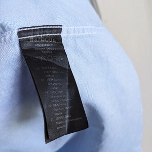 May include: A light blue shirt with a black tag attached. The tag has the brand name "Barbour" printed on it, along with other text. The shirt is made of cotton.