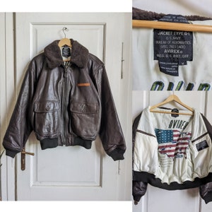 May include: A brown leather jacket with a shearling collar, two front pockets, and a black ribbed hem and cuffs. The jacket has a patch on the chest and the lining features a map of the United States and the text "AVIREX".