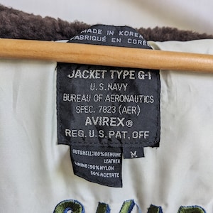 May include: Close-up of the inside of a vintage Avirex G-1 jacket. The black tag reads "JACKET TYPE G-1 U.S. NAVY BUREAU OF AERONAUTICS AVIREX&reg;". The jacket is made of genuine leather and has a nylon and acetate lining. The jacket is size M.