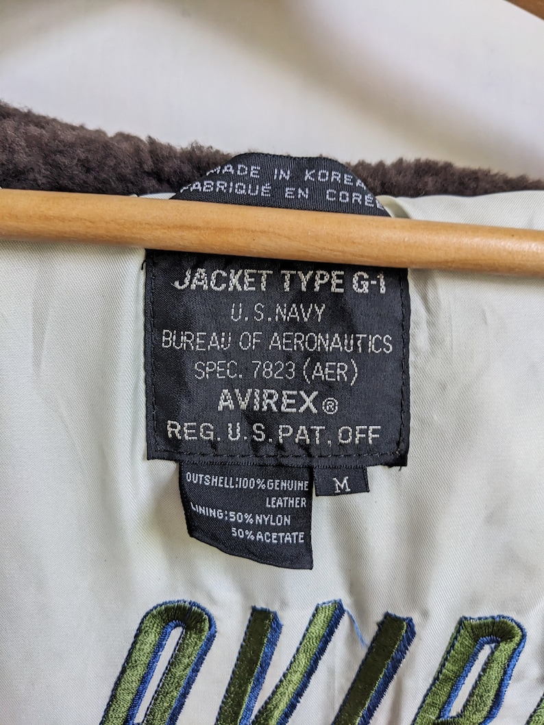 May include: Close-up of the inside of a vintage Avirex G-1 jacket. The black tag reads "JACKET TYPE G-1 U.S. NAVY BUREAU OF AERONAUTICS AVIREX&reg;". The jacket is made of genuine leather and has a nylon and acetate lining. The jacket is size M.