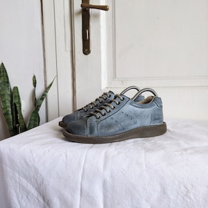 Puede incluir: Un par de zapatillas de cuero azul con suelas y cordones marrones. Los zapatos se exhiben sobre una superficie blanca, con una puerta blanca y una planta en el fondo. Las zapatillas tienen un diseño clásico y discreto.