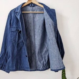 May include: A blue denim jacket with a button closure and two pockets. The jacket is unbuttoned and hanging on a wooden hanger.