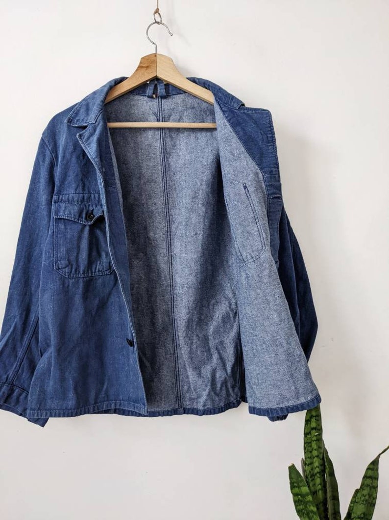 May include: A blue denim jacket with a button closure and two pockets. The jacket is unbuttoned and hanging on a wooden hanger.