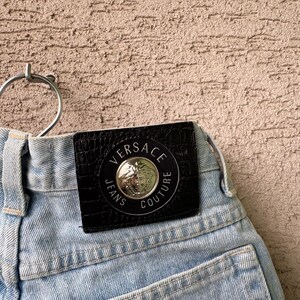 K&ouml;nnte beinhalten: Hellblaue Jeans aus Denim mit einem schwarzen Lederpatch mit dem Versace Jeans Couture-Logo. Der Patch ist kreisf&ouml;rmig mit silberner Schrift und einem zentralen Emblem. Die Jeans haben ein klassisches F&uuml;nf-Taschen-Design.
