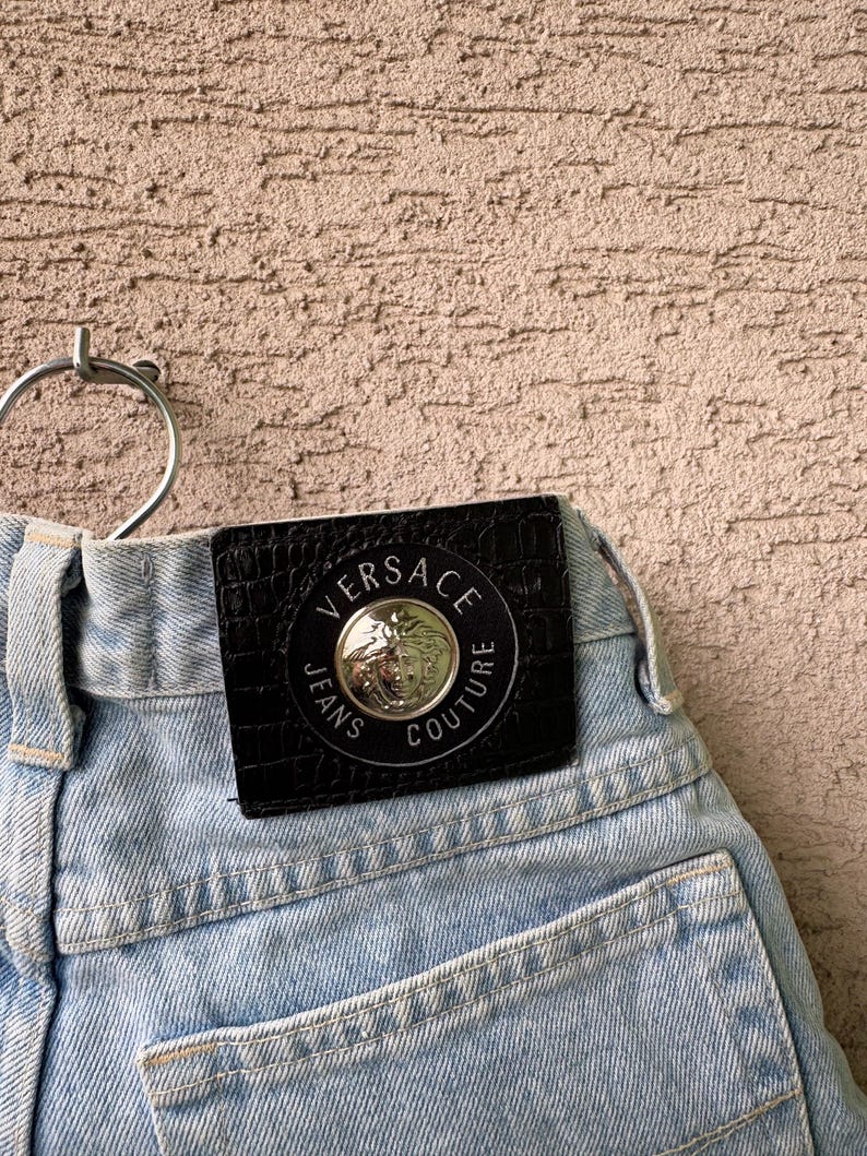 K&ouml;nnte beinhalten: Hellblaue Jeans aus Denim mit einem schwarzen Lederpatch mit dem Versace Jeans Couture-Logo. Der Patch ist kreisf&ouml;rmig mit silberner Schrift und einem zentralen Emblem. Die Jeans haben ein klassisches F&uuml;nf-Taschen-Design.
