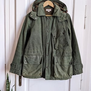 Pode incluir: Uma jaqueta verde com capuz, fecho de botões e vários bolsos. A jaqueta tem uma etiqueta de couro marrom com o texto "Baracuta" nela.
