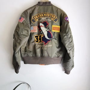 Puede incluir: Chaqueta bomber verde oliva con parches bordados. La parte trasera presenta un gr&aacute;fico de tibur&oacute;n, el texto "SHARK MOUTHS 388th TACTICAL FIGHTER WING" y el n&uacute;mero "34". Incluye banderas americanas y otras. La chaqueta est&aacute; colgada en una pared blanca.