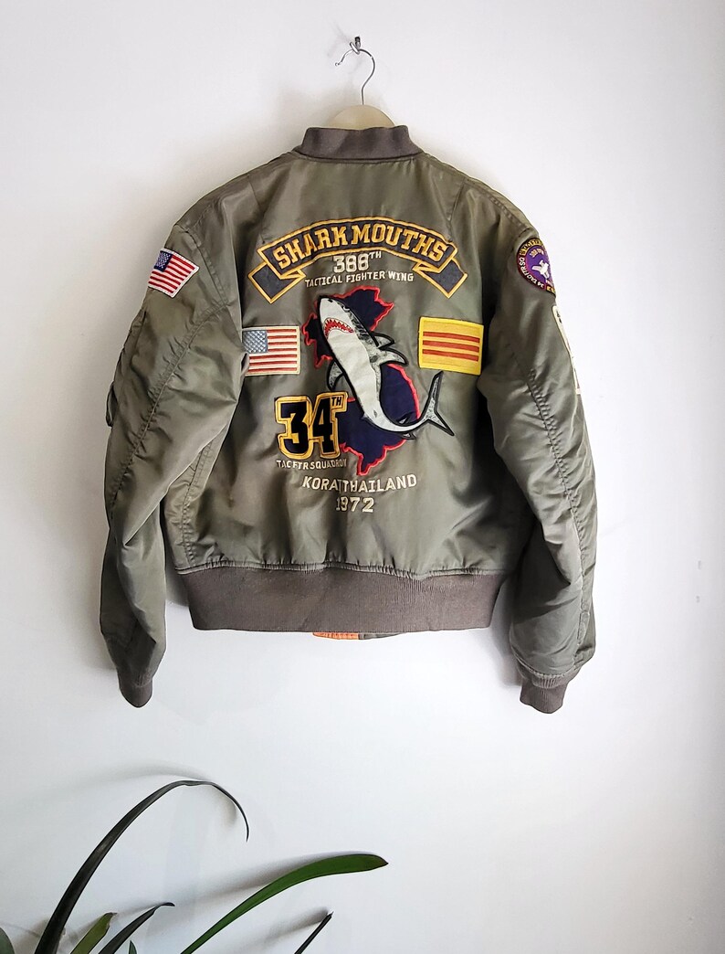 Puede incluir: Chaqueta bomber verde oliva con parches bordados. La parte trasera presenta un gr&aacute;fico de tibur&oacute;n, el texto "SHARK MOUTHS 388th TACTICAL FIGHTER WING" y el n&uacute;mero "34". Incluye banderas americanas y otras. La chaqueta est&aacute; colgada en una pared blanca.