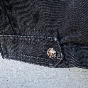 Puede incluir: Primer plano de un detalle de chaqueta vaquera negra. La chaqueta presenta un bot&oacute;n plateado con un dise&ntilde;o en relieve. La tela tiene un aspecto texturizado y las costuras son visibles.