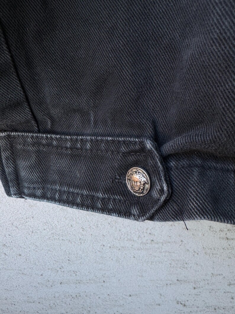 Puede incluir: Primer plano de un detalle de chaqueta vaquera negra. La chaqueta presenta un bot&oacute;n plateado con un dise&ntilde;o en relieve. La tela tiene un aspecto texturizado y las costuras son visibles.