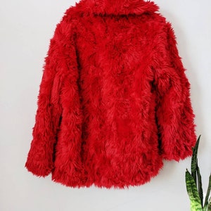 Puede incluir: Una chaqueta de piel sint&eacute;tica roja con cuello. La chaqueta est&aacute; colgada de una percha contra una pared blanca.