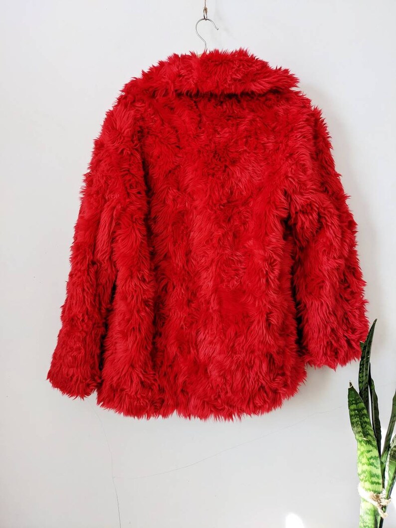 Puede incluir: Una chaqueta de piel sint&eacute;tica roja con cuello. La chaqueta est&aacute; colgada de una percha contra una pared blanca.