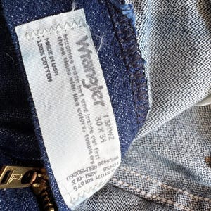 Op de afbeelding: Close-up van een blauwe Wrangler jeans. Het label luidt "Wrangler 13MWZ 30 x 34" en "Made in USA 100% Cotton". Een gouden rits is zichtbaar. De jeans is gemaakt van een duurzame denim stof.