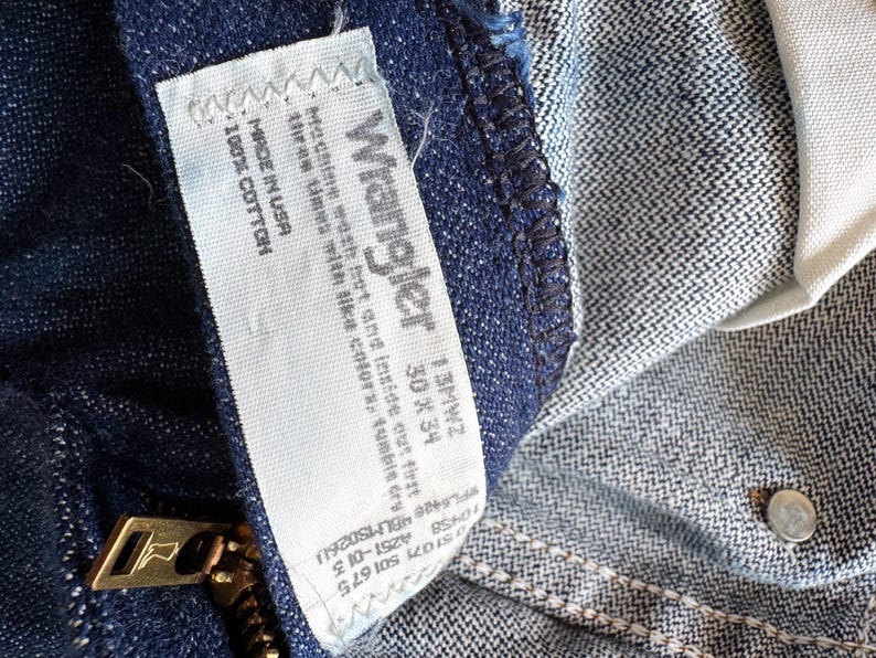 Op de afbeelding: Close-up van een blauwe Wrangler jeans. Het label luidt "Wrangler 13MWZ 30 x 34" en "Made in USA 100% Cotton". Een gouden rits is zichtbaar. De jeans is gemaakt van een duurzame denim stof.