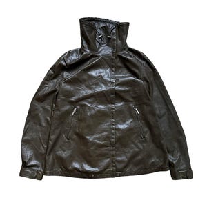 May include: A dark brown jacket with a high funnel neck, featuring a front zipper closure, two side pockets with silver-toned zippers, and adjustable cuffs. The material has a smooth, slightly reflective finish with subtle creasing throughout.