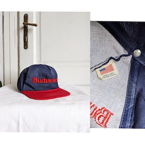 Puede incluir: Una gorra de béisbol azul y roja con la palabra "Budweiser" bordada en rojo en la parte delantera. La gorra tiene un forro de malla blanca y una etiqueta que dice "Made in U.S.A. One Size Fits All".
