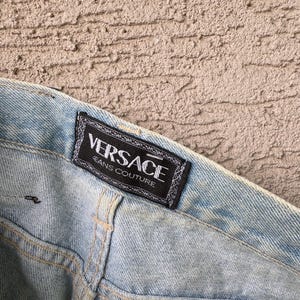 K&ouml;nnte beinhalten: Hellblaue Jeans mit einem sichtbaren "VERSACE JEANS COUTURE" Etikett. Das Etikett ist schwarz mit wei&szlig;em Text und einem dekorativen Rand. Die Jeans hat sichtbare N&auml;hte und ein verwaschenes Aussehen.