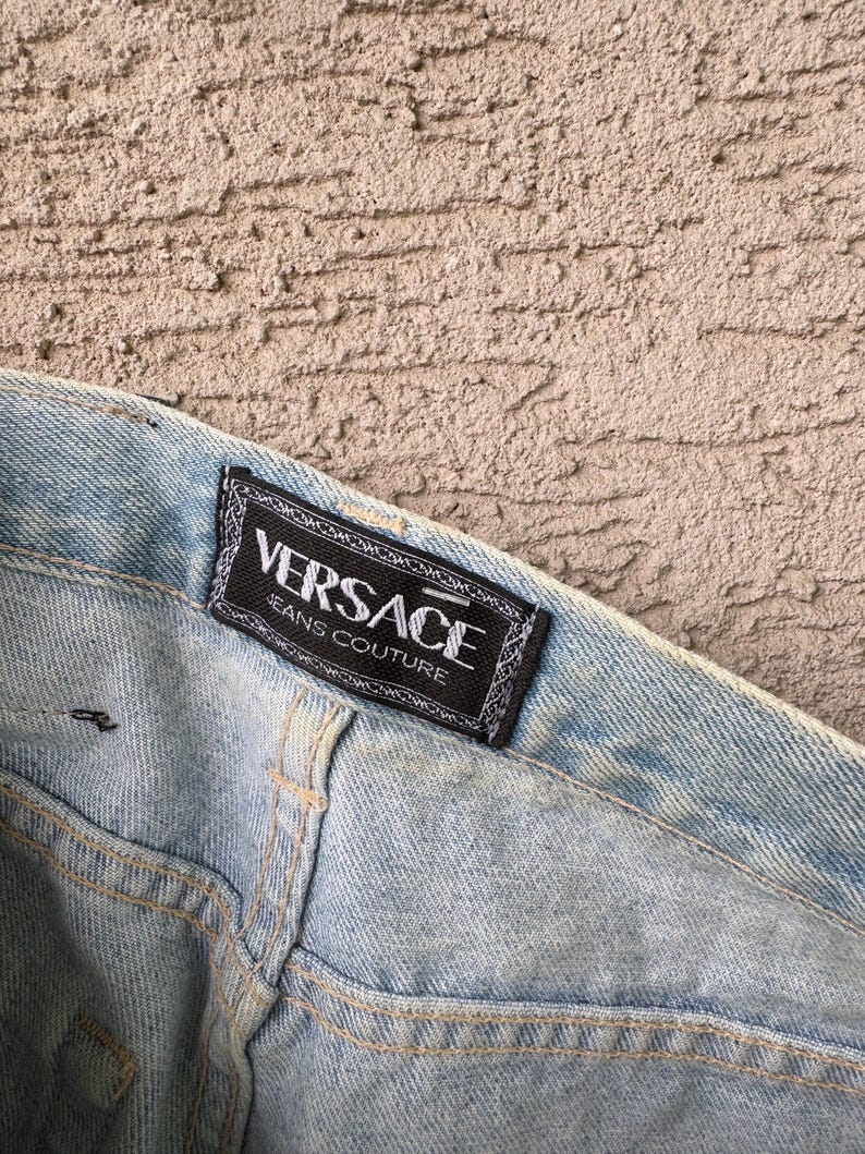 K&ouml;nnte beinhalten: Hellblaue Jeans mit einem sichtbaren "VERSACE JEANS COUTURE" Etikett. Das Etikett ist schwarz mit wei&szlig;em Text und einem dekorativen Rand. Die Jeans hat sichtbare N&auml;hte und ein verwaschenes Aussehen.