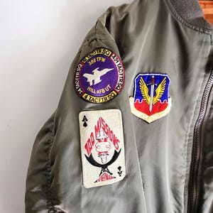 Puede incluir: Chaqueta bomber verde oliva con m&uacute;ltiples parches bordados. Los parches incluyen un avi&oacute;n morado y blanco, un escudo con una espada y alas, y un dise&ntilde;o de carta de juego con el texto "100 MISSIONS". La chaqueta tiene cuello y pu&ntilde;os acanalados.